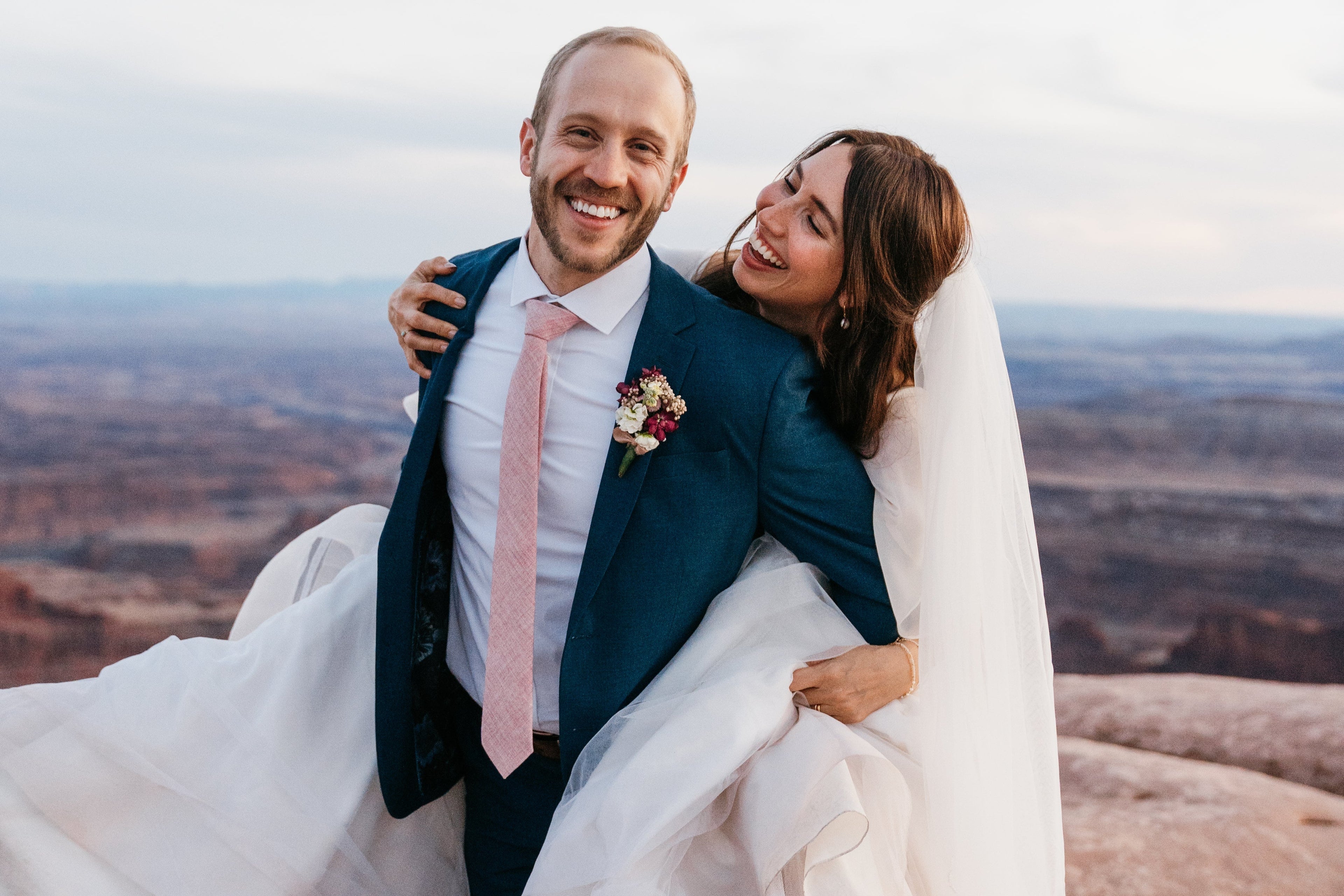 Man in a pink tie holding a woman in a wedding dress with a scenic background