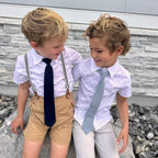 Two young boys wearing ties and suspenders standing outdoors against a stone wall.