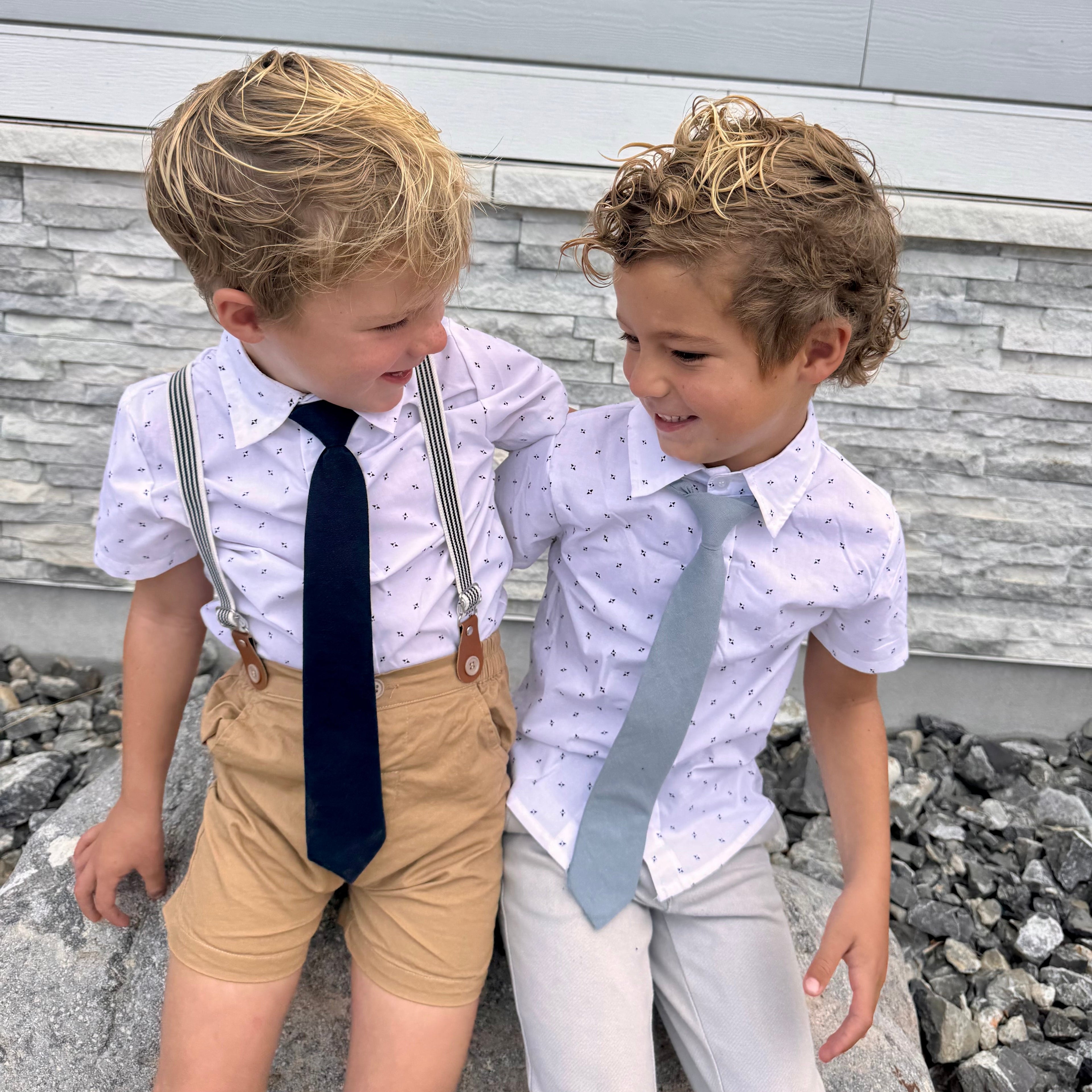 Two young boys wearing ties and suspenders standing outdoors against a stone wall.
